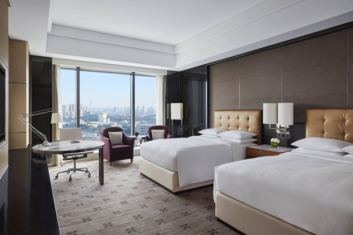 Habitación de hotel con dos camas, un escritorio, una silla y un amplio ventanal con vista a la ciudad.