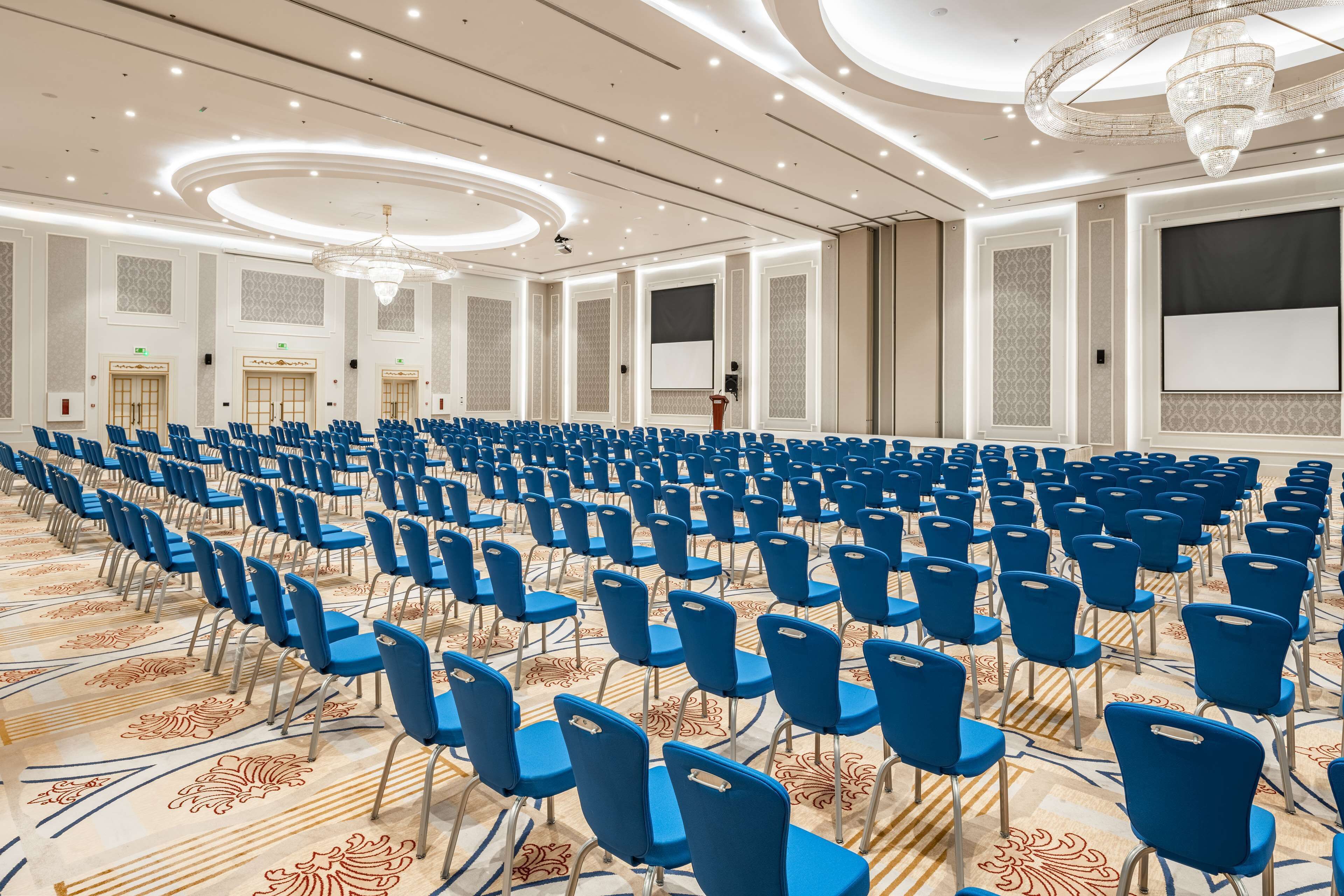 A conference room with rows of blue chairs, a large screen, and a podium.