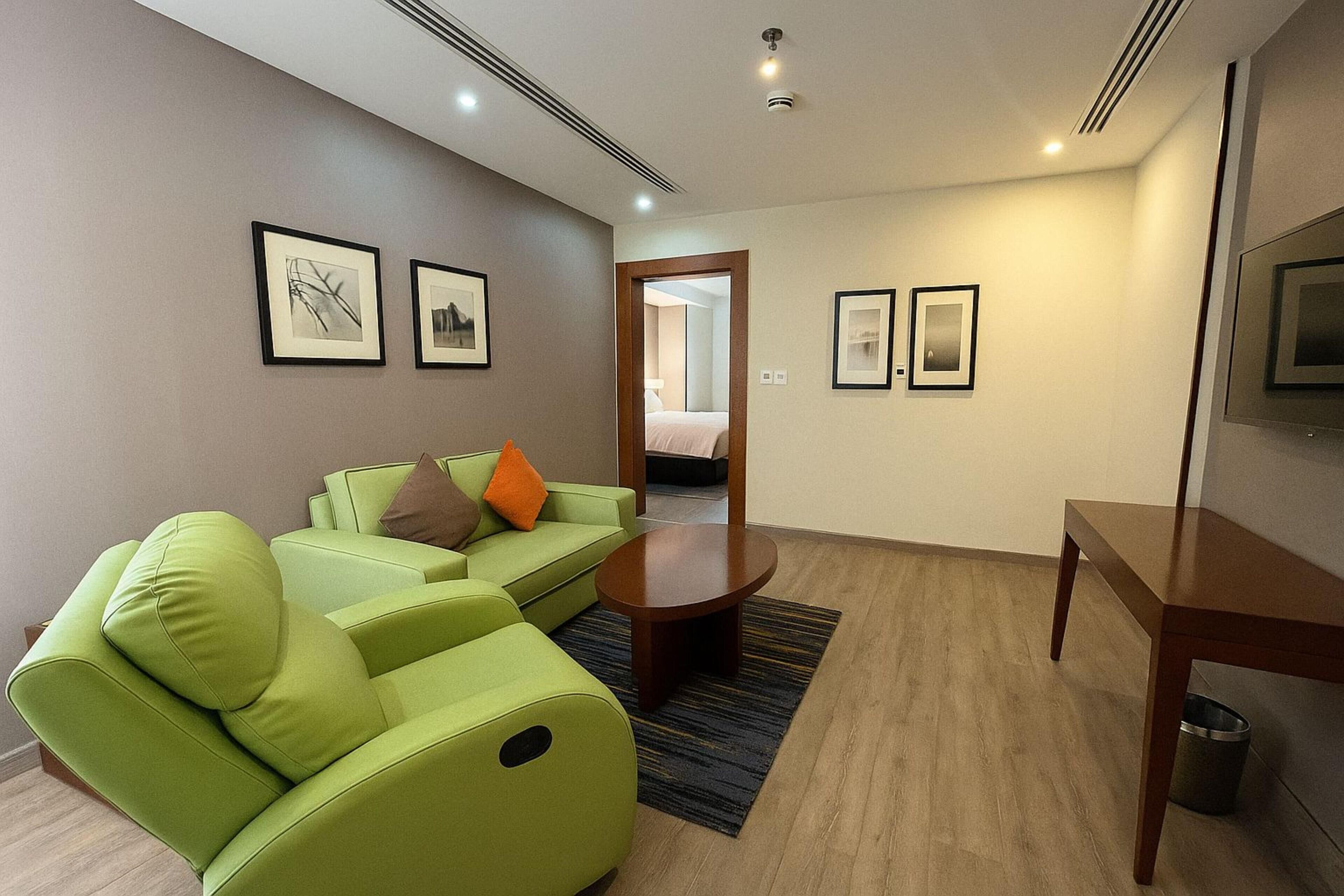 Een moderne hotelkamer met een groene bank, een houten bijzettafeltje en kunstwerken in lijsten aan de wanden.