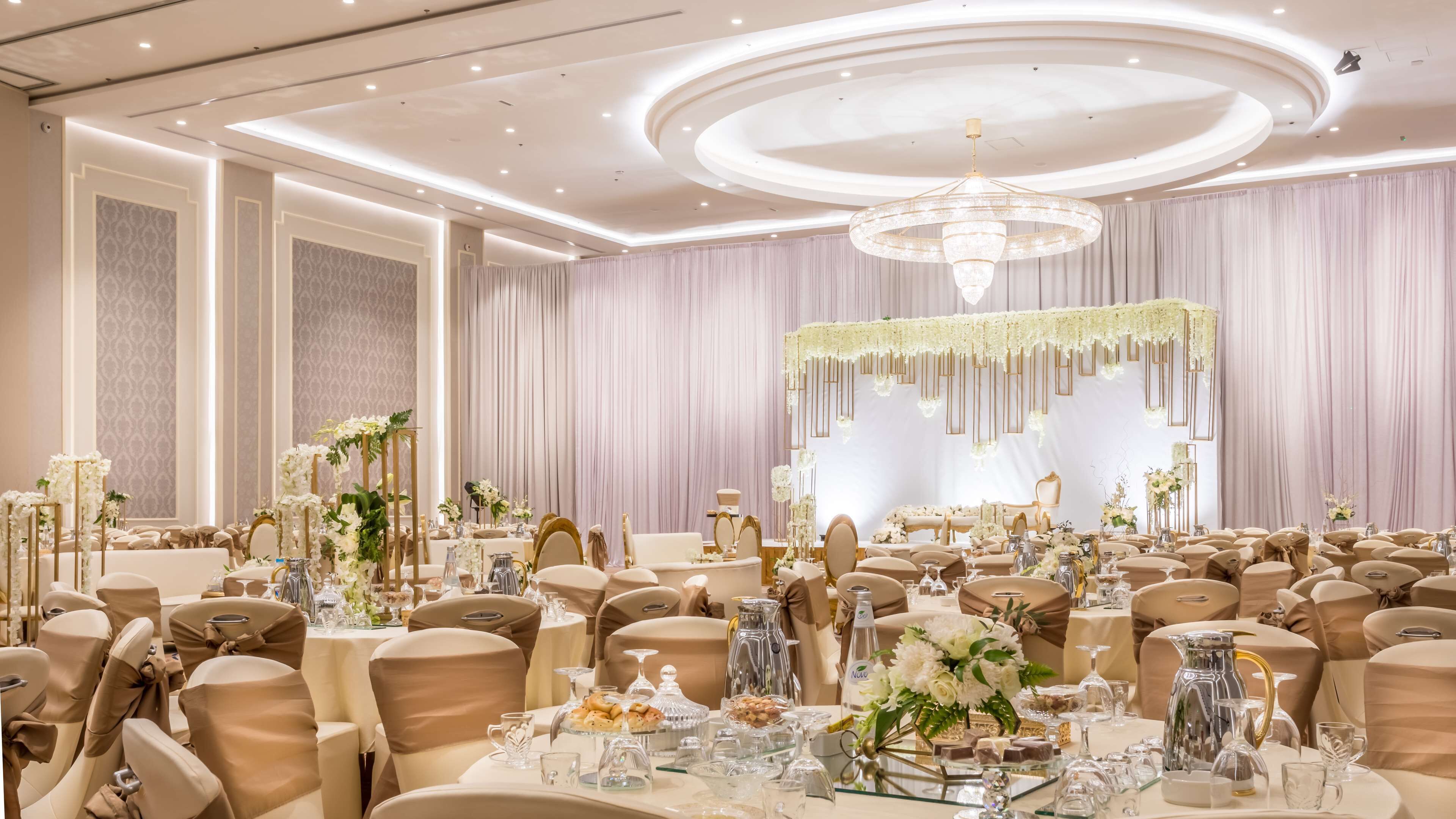 Une salle de banquet spacieuse, dotée de tables rondes, d’élégants lustres et de décorations florales.