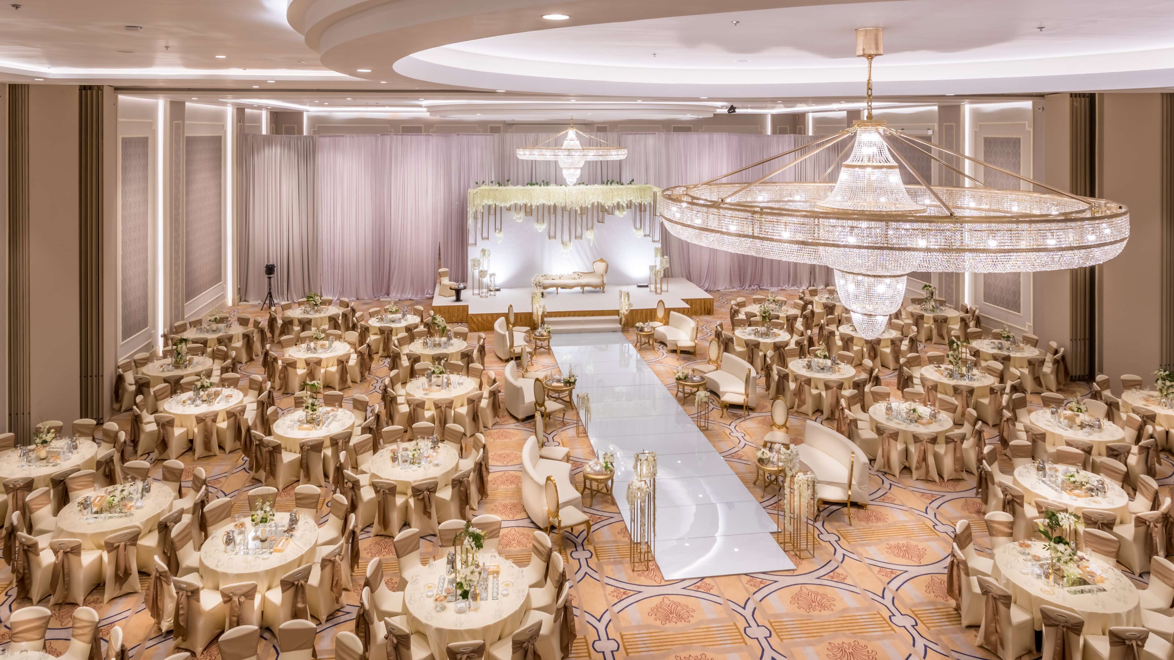 A banquet hall with round tables, a central aisle, and a stage with a chandelier.