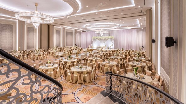 A spacious banquet hall with round tables, elegant chandeliers, and a grand staircase.