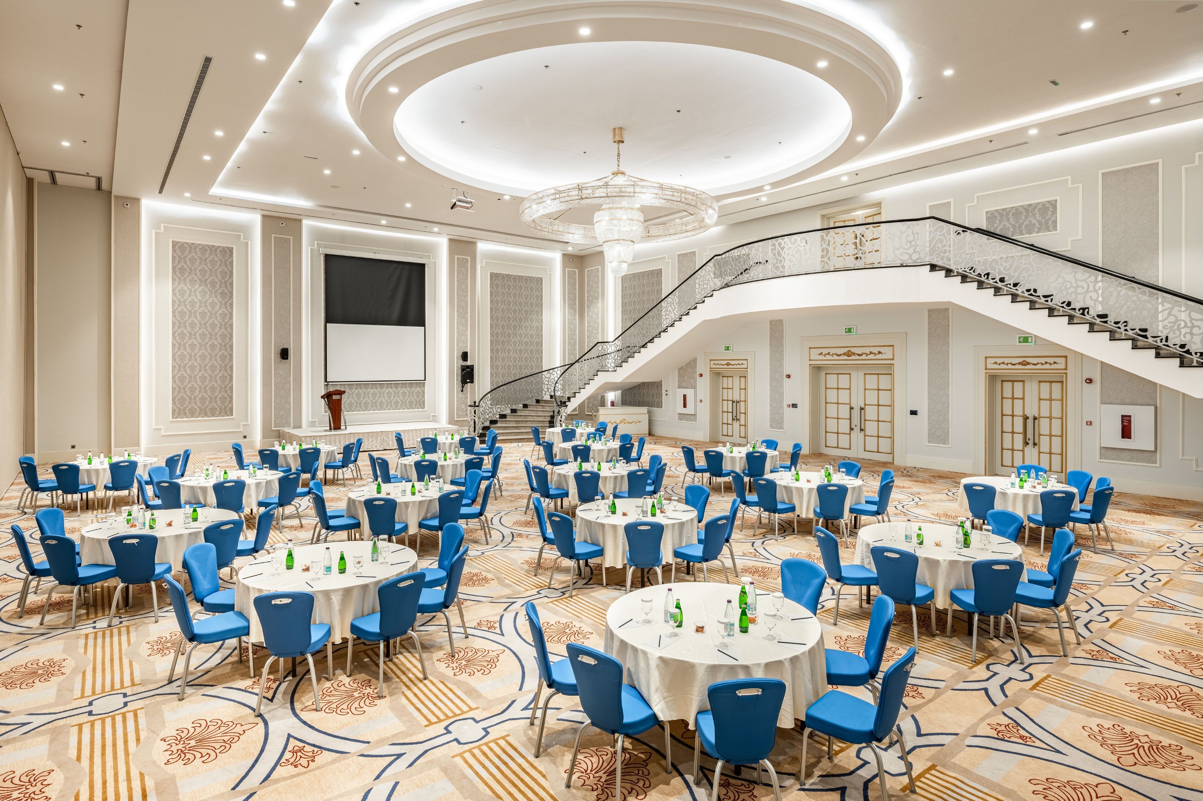 Une salle de conférence spacieuse, dotée de tables rondes, de chaises bleues et d’un magnifique escalier.