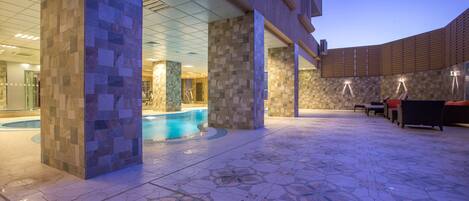 A pool area with tiled flooring and a pool, surrounded by pillars and a covered seating area with chairs.