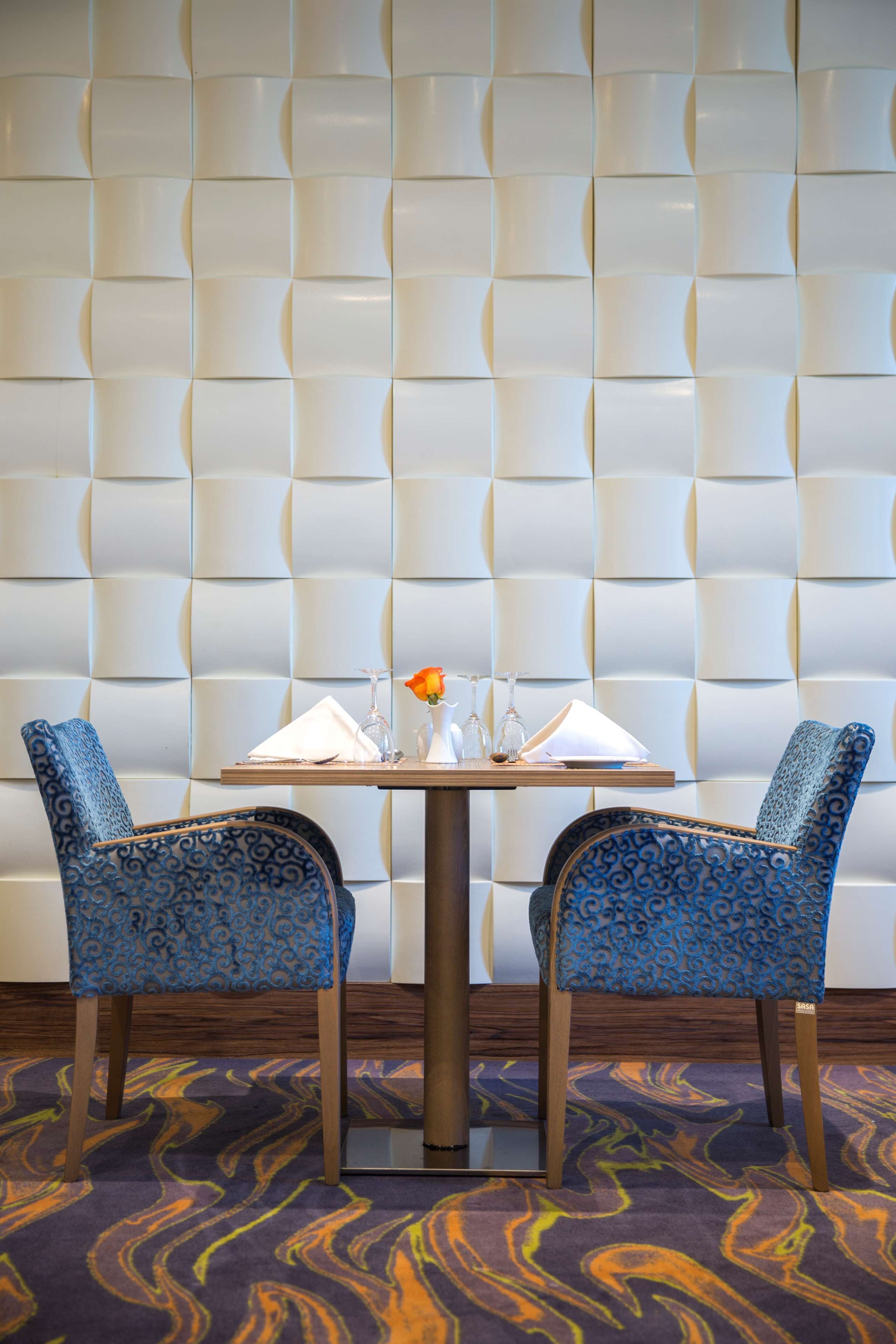 A dining area with a tufted wall, two patterned chairs, a table set with a white napkin, a glass vase with a single flower, and a tray with glasses and a bottle.