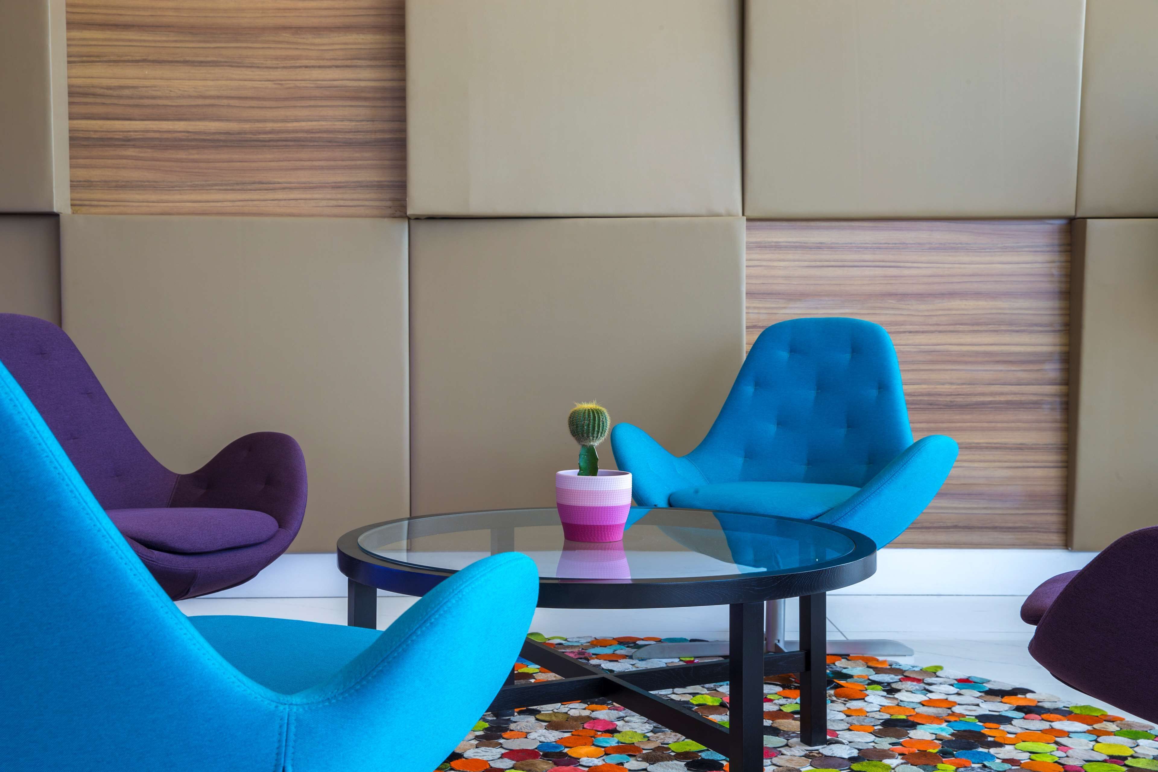 A modern room with a glass table, two blue chairs, a purple chair, and a colorful rug.