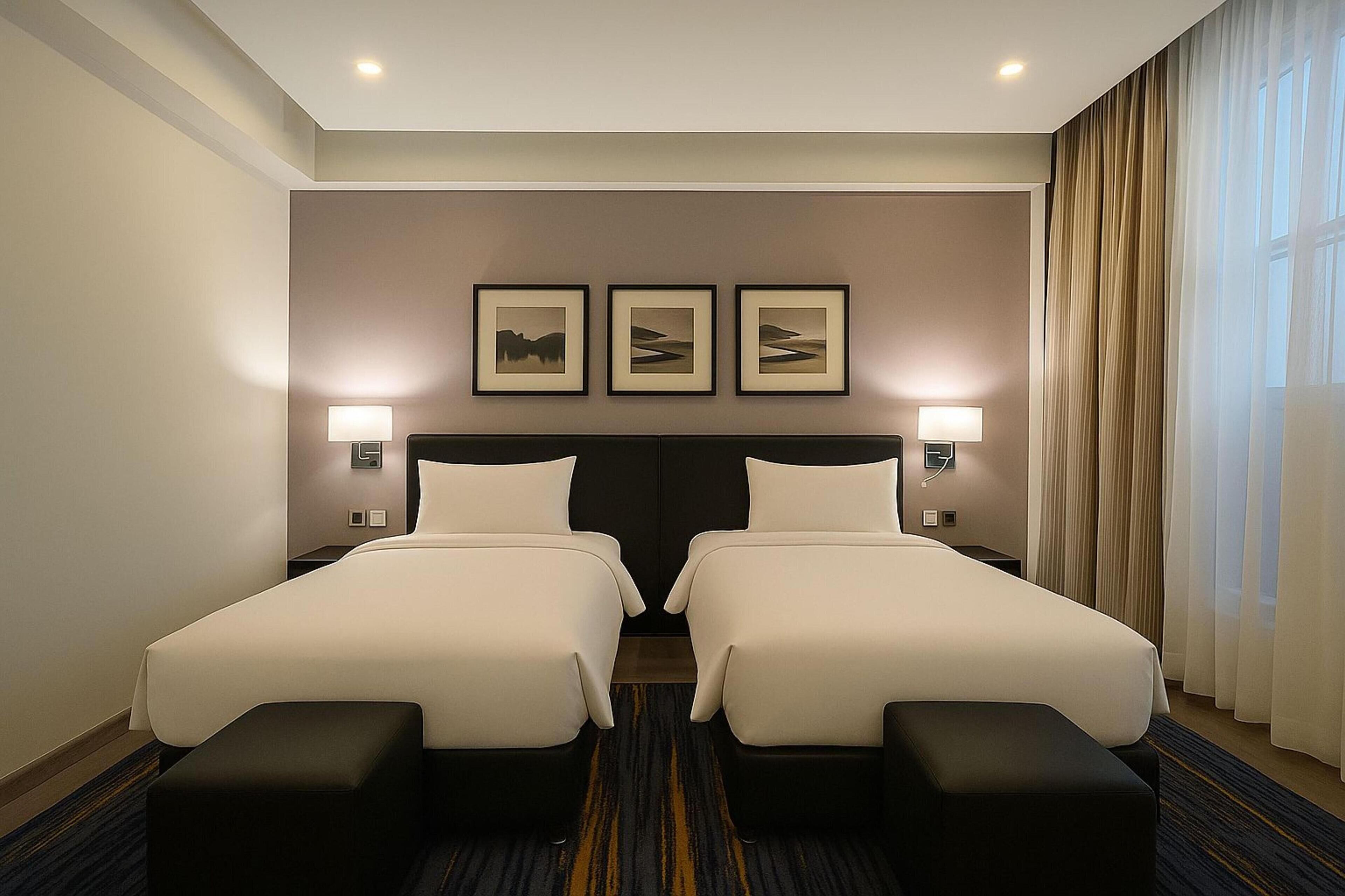 Ett hotellrum med två sängar, ett stort fönster med gardiner och tre inramade tavlor på väggen.