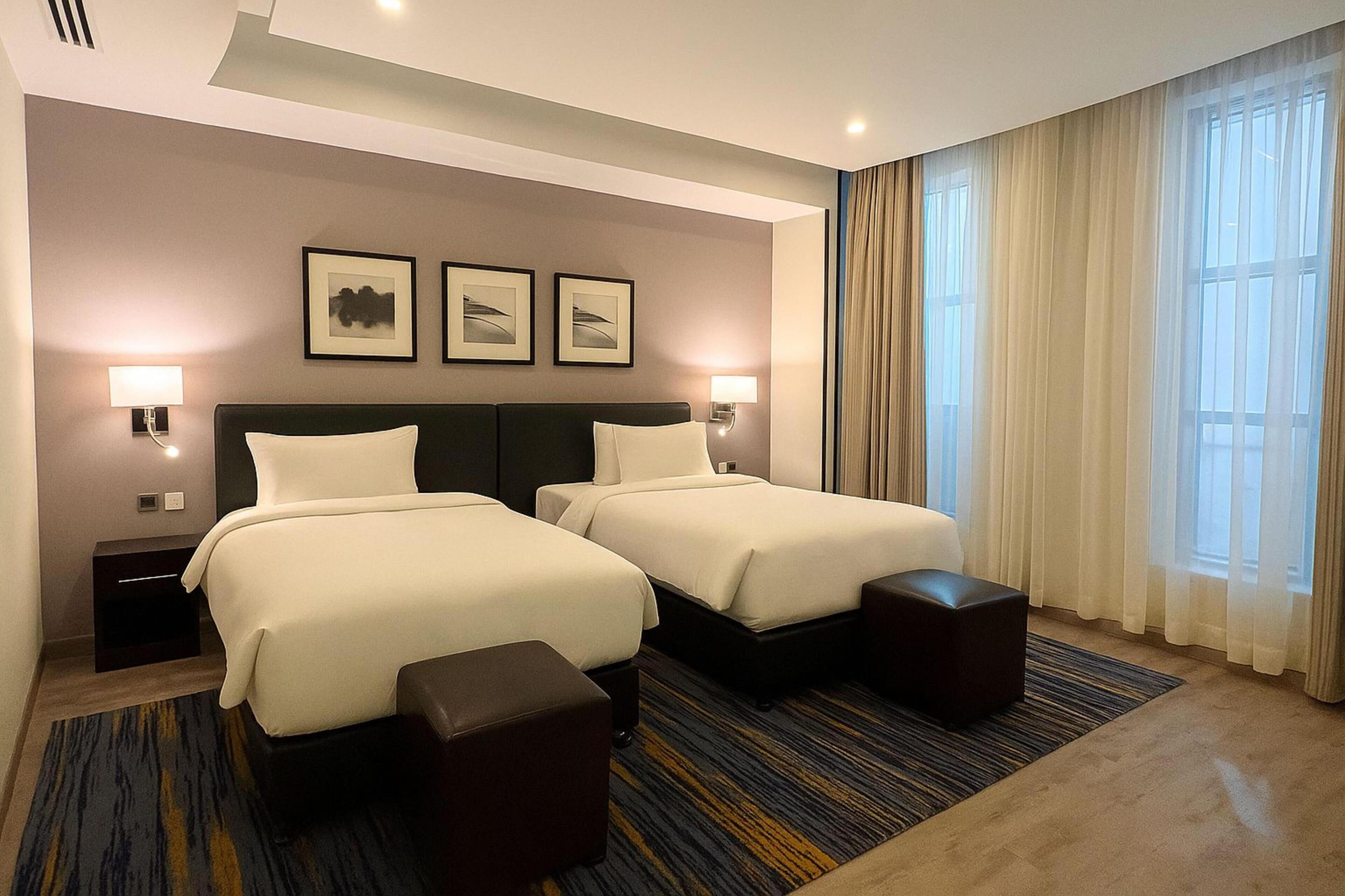 A hotel room with two beds, a large window with curtains, and framed artwork on the wall.