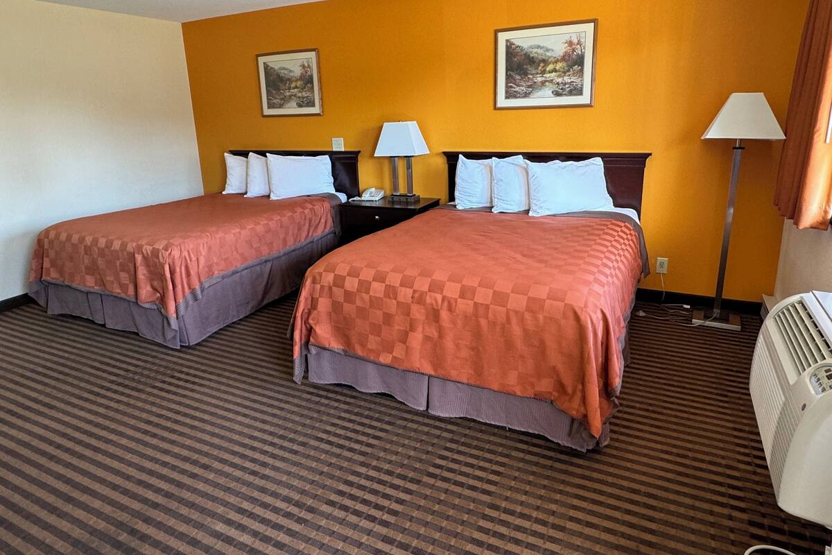 A hotel room with two beds, a nightstand with a lamp, a wall-mounted heater, and two framed pictures on the wall.