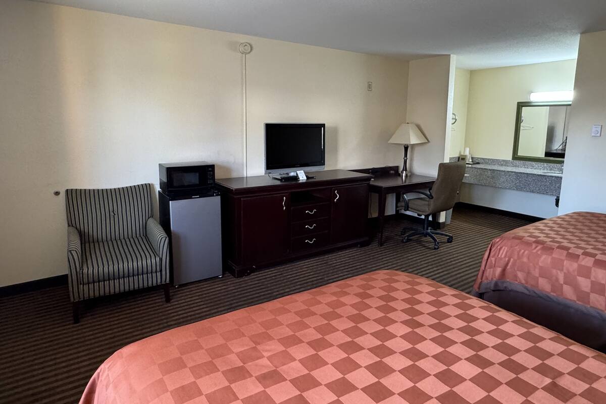 A hotel room with two beds, a desk with a television, a refrigerator, a striped chair, and a lamp.
