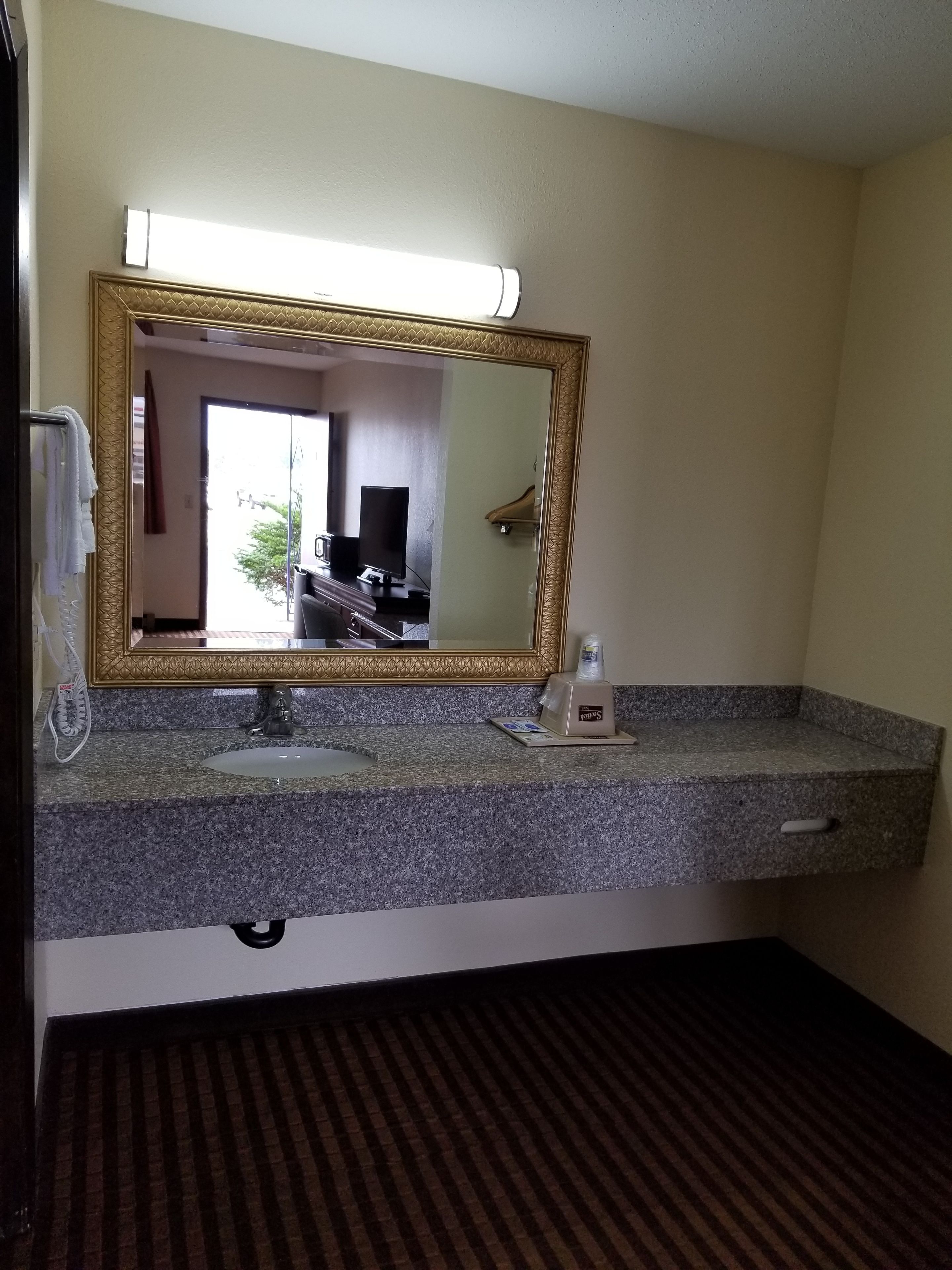 A hotel bathroom with a granite countertop, a sink, a mirror with a gold frame, a towel rack, and a shower area visible through the glass door.