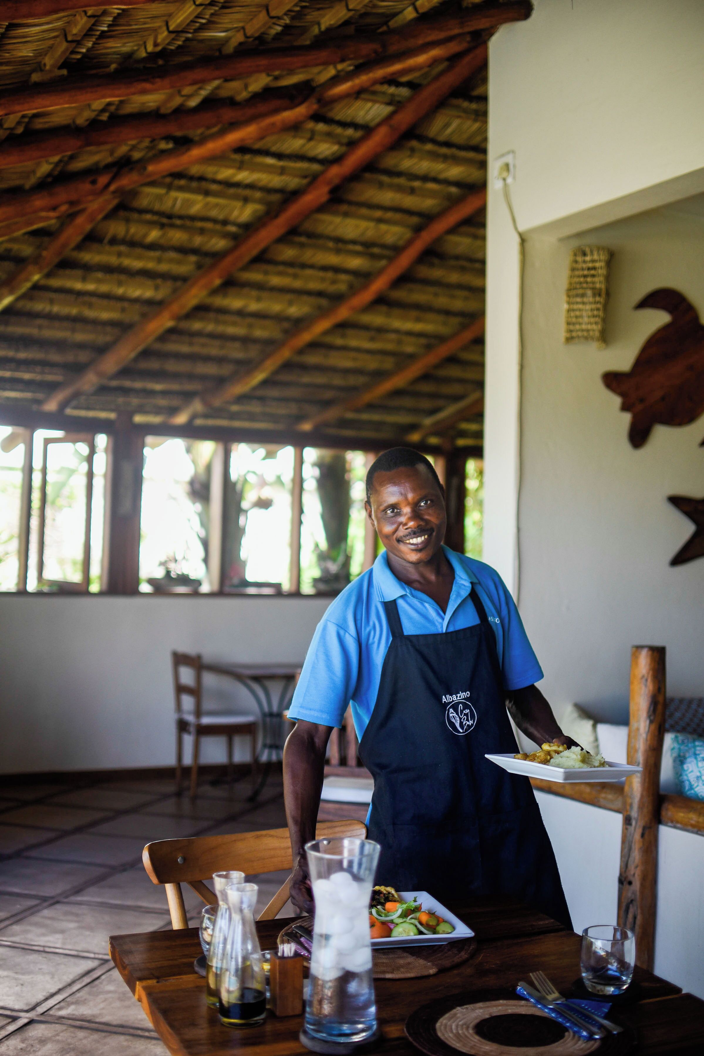 A person in a blue shirt and apron standing in a room with a thatched roof, wooden furniture, and a dining table set with plates, glasses, and cutlery.