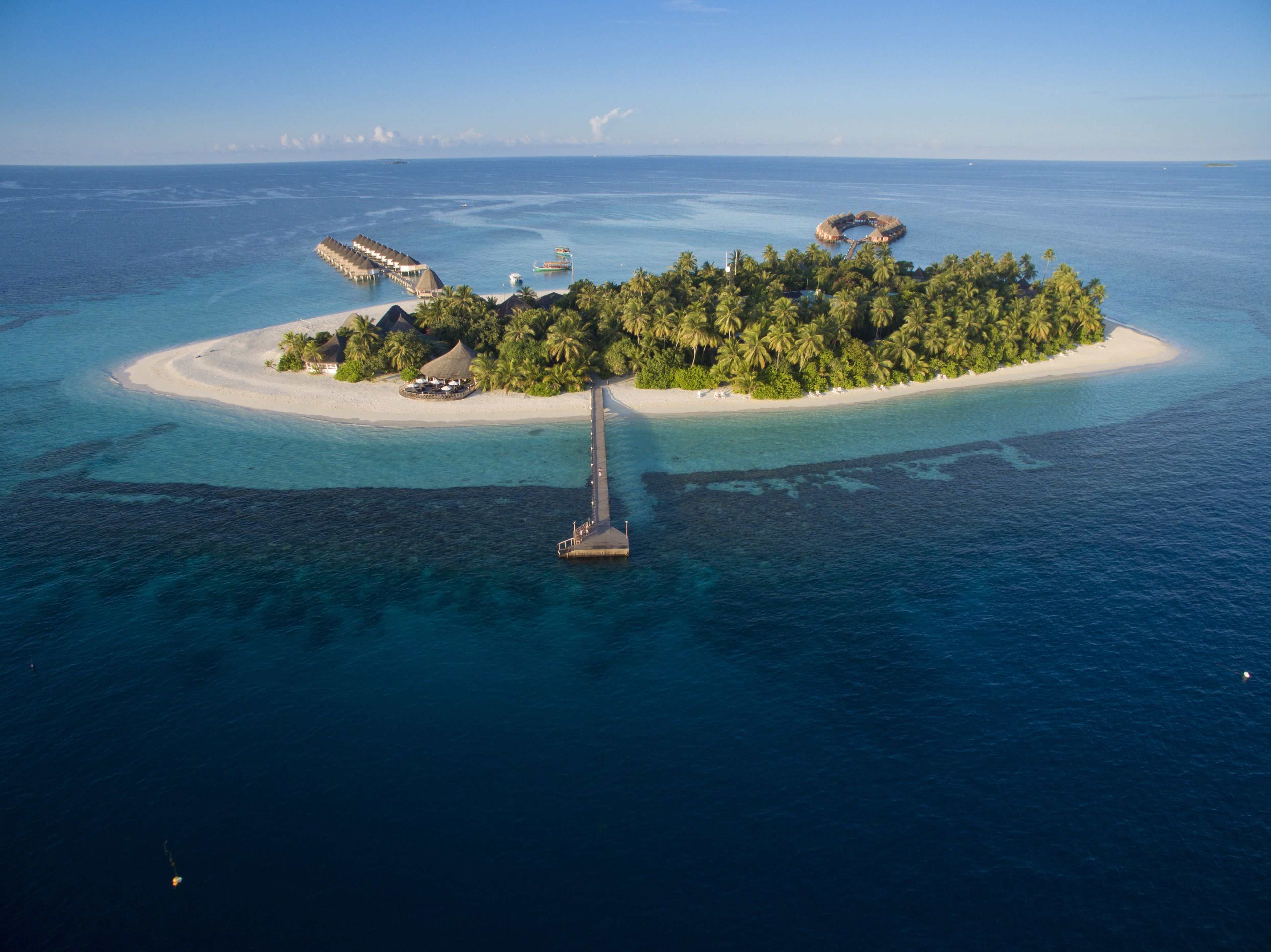 An aerial view of a tropical island with a pier leading to a resort area, surrounded by clear blue waters and palm trees.