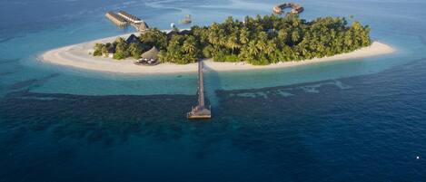 An aerial view of a tropical island with a pier leading to a resort area, surrounded by clear blue waters and palm trees.
