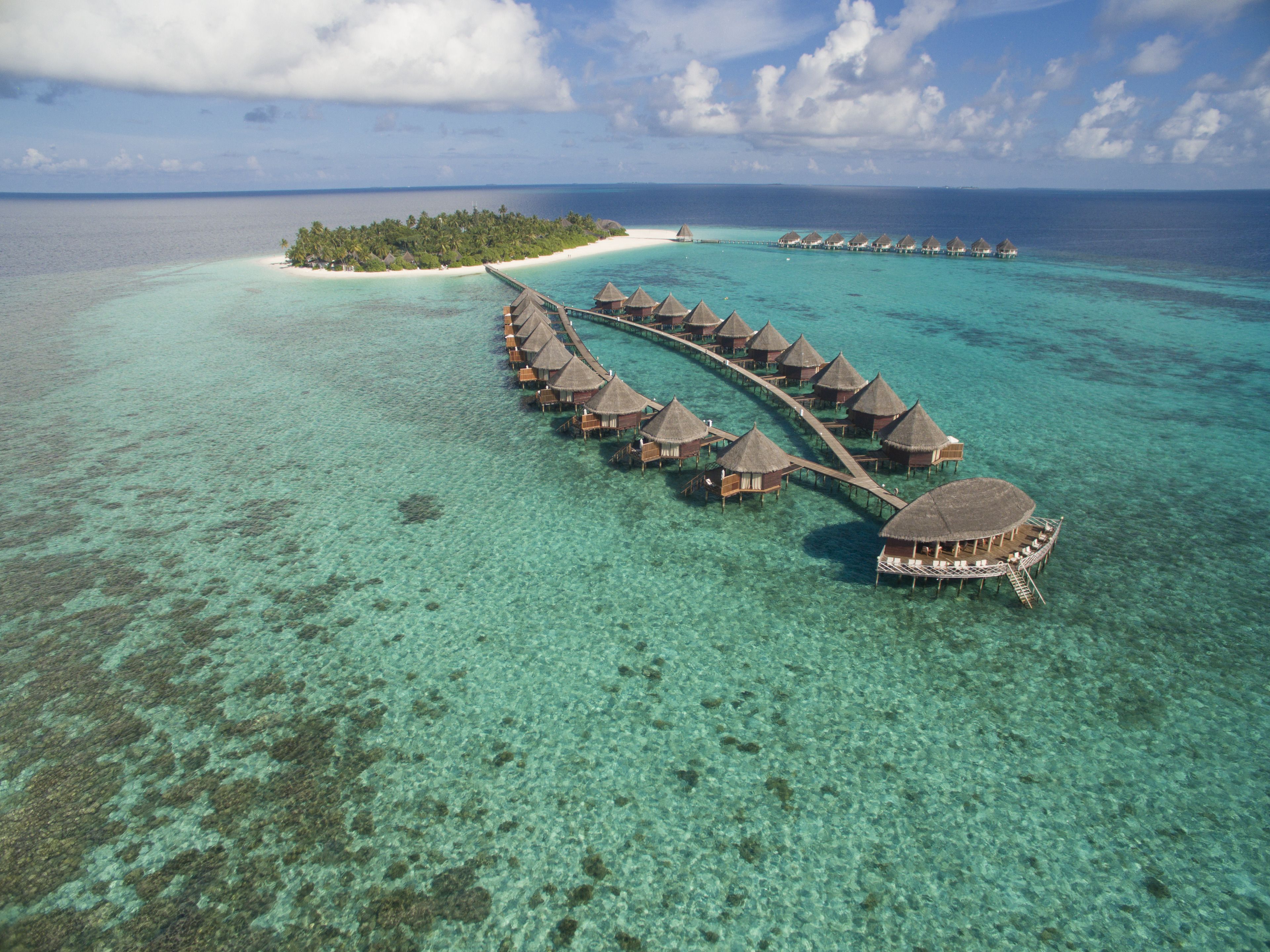 An aerial view of overwater bungalows connected by a walkway, surrounded by clear turquoise waters and a small island with palm trees.