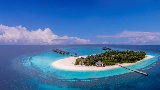 Vista aérea de una isla tropical con bungalows sobre el agua, una playa de arena blanca y aguas turquesas cristalinas.