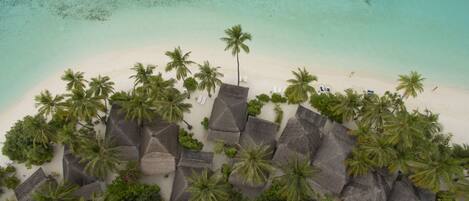 Vista aérea de un resort en la playa con cabañas de techo de paja, palmeras y aguas turquesas cristalinas.