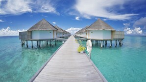 Un muelle de madera que conduce a bungalós sobre el agua con techos de paja, agua turquesa cristalina y un cielo azul brillante.