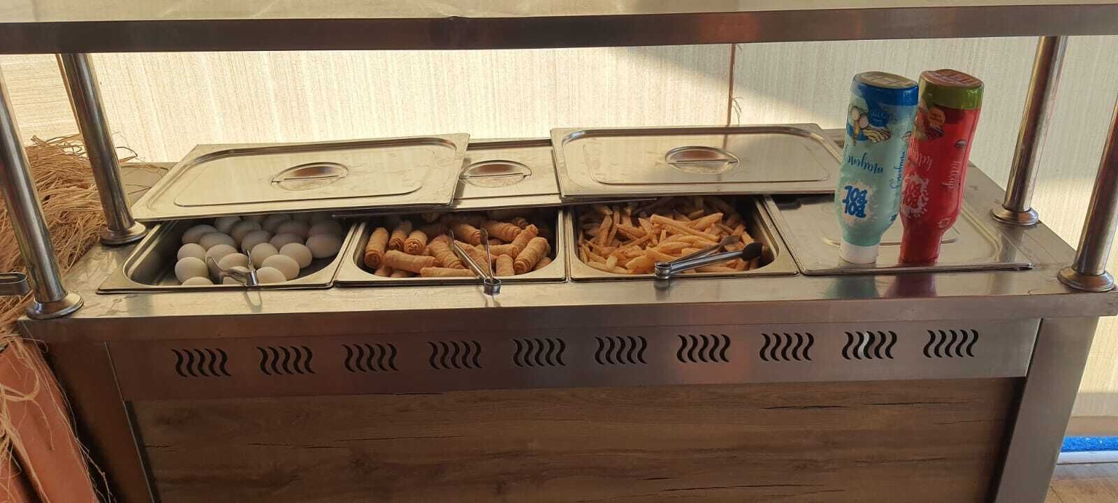 Un bufé con bandejas de huevos, buñuelos fritos y papas fritas, y dos dispensadores de bebidas.