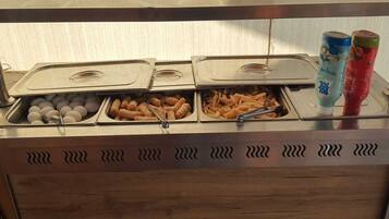 Un bufé con bandejas de huevos, buñuelos fritos y papas fritas, y dos dispensadores de bebidas.
