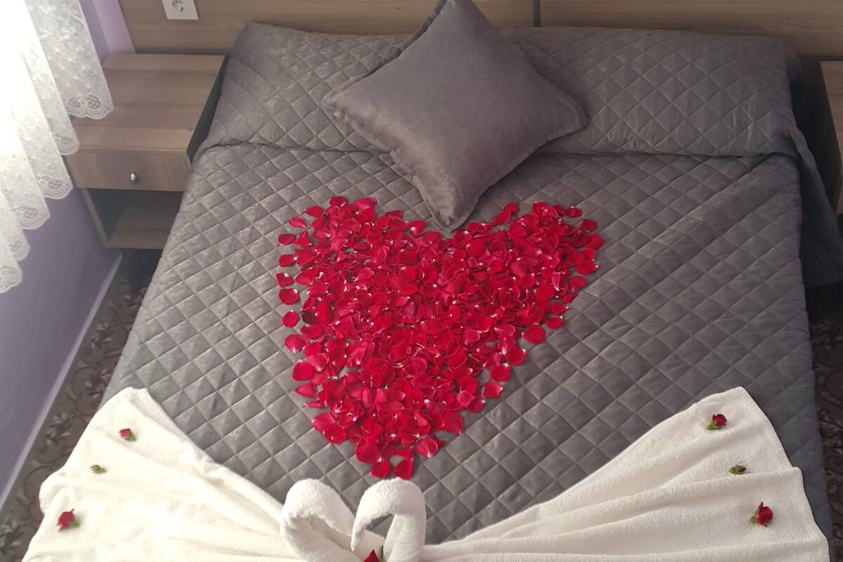 Un lit recouvert d’une courtepointe grise matelassée, un oreiller et une composition en forme de cœur faite de pétales de roses rouges, disposée sur le lit.