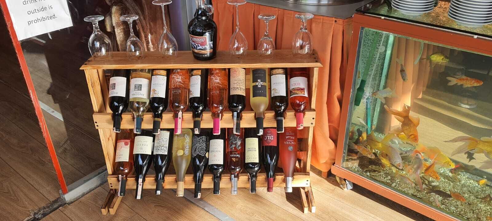 Una estantería con varias botellas y copas de vino, un acuario con peces dorados, y un letrero que prohíbe el consumo de bebidas alcohólicas fuera del establecimiento.