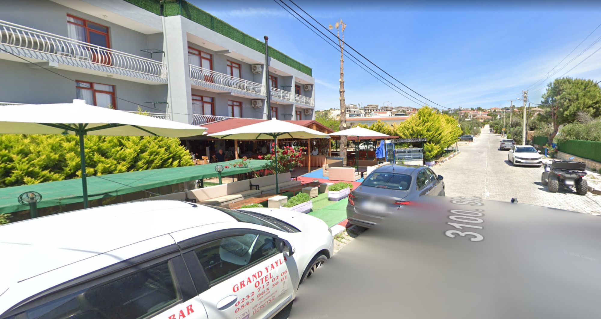 Vista de la calle con un coche blanco estacionado al lado, un edificio con balcones y zonas de estar al aire libre.