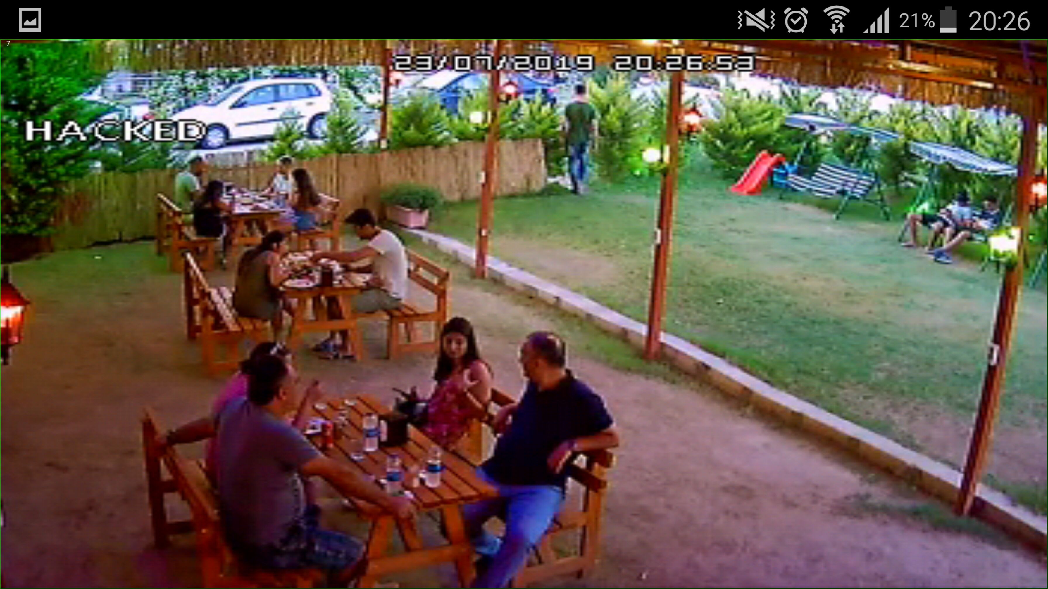 An outdoor seating area with wooden tables and chairs, surrounded by greenery and a slide.