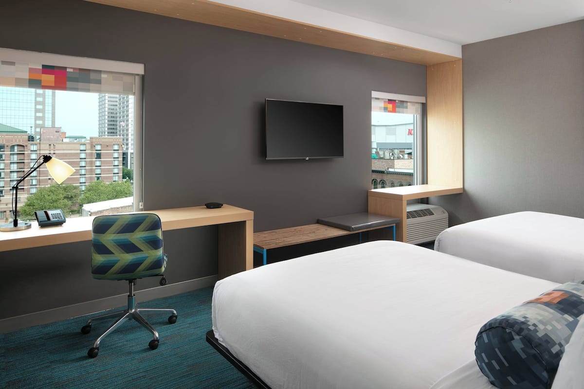 A hotel room with a bed, desk, chair, TV, and windows with city views.