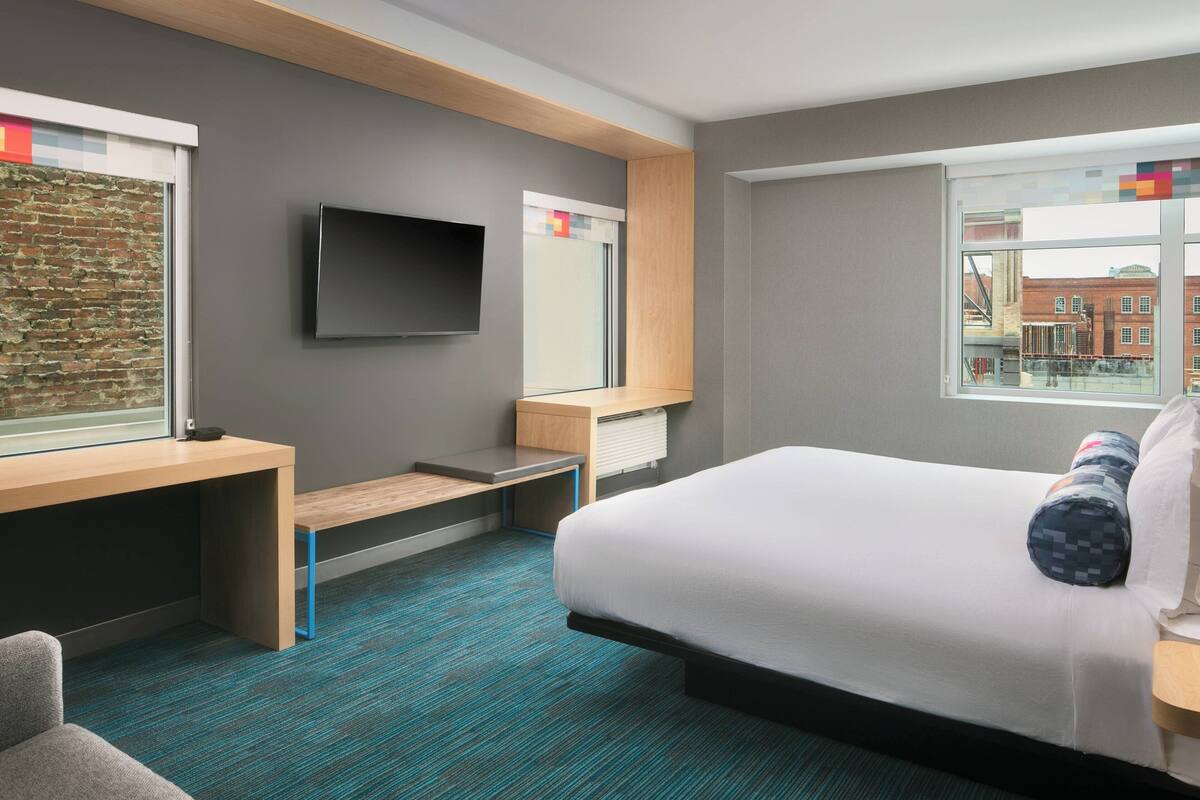 A hotel room with a bed, a desk, a television, and a window with a city view.