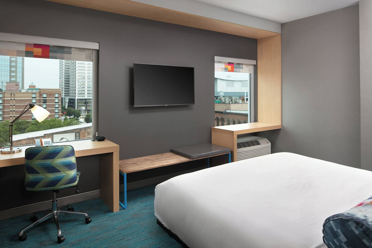A modern hotel room with a large bed, a desk with a chair, a flat-screen TV, and a window with a city view.
