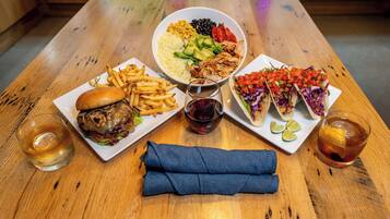 Una mesa de madera con diversos alimentos, incluyendo una hamburguesa, papas fritas, una ensalada y tacos, junto con dos vasos de una bebida oscura.