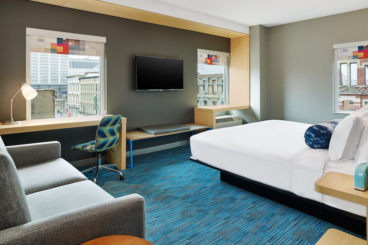 A hotel room with a bed, a desk, a chair, a TV, and windows with city views.