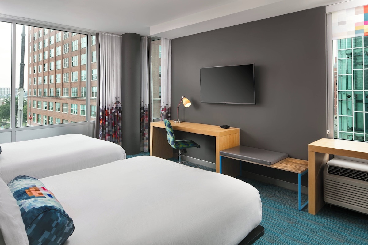 Habitación de hotel con dos camas, un escritorio, una silla, un televisor y una ventana con vista a la ciudad.