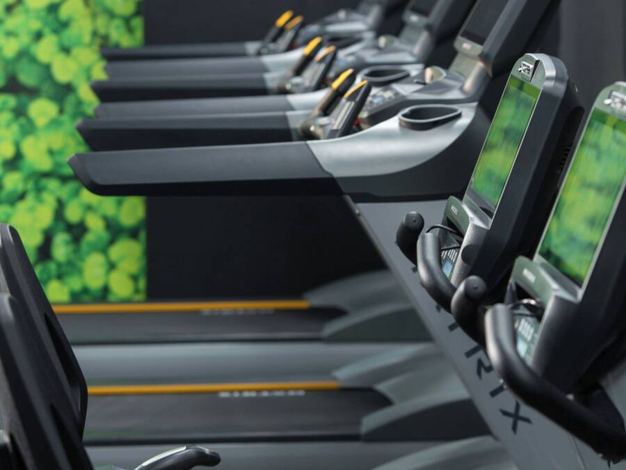 A row of treadmills with digital screens, set against a green leafy wall.