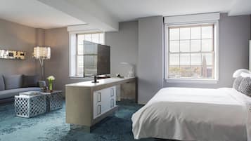 A modern hotel room with a large bed, a desk with a flat-screen TV, a sofa, and a large window offering a city view.