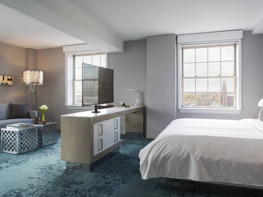 A modern hotel room with a large bed, a desk with a flat-screen TV, a sofa, and a large window offering a city view.