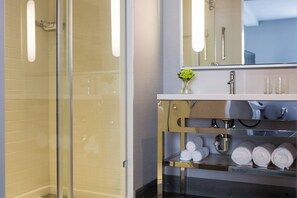 A modern bathroom with a glass shower enclosure, a white sink with a vase of flowers, and neatly rolled towels on a shelf.