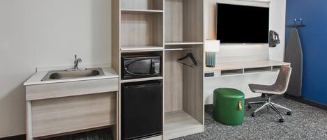 Una habitación compacta con escritorio integrado, microondas, lavamanos, una silla y un cubo de basura verde.