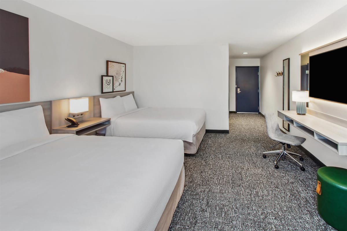 A hotel room with two beds, a desk with a chair, a TV, and a green trash bin.