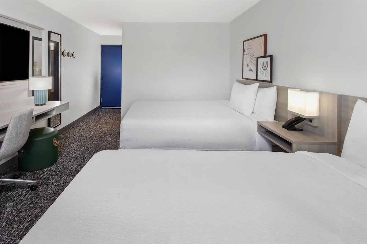 A hotel room with two beds, a desk with a chair, a trash bin, and a blue door.