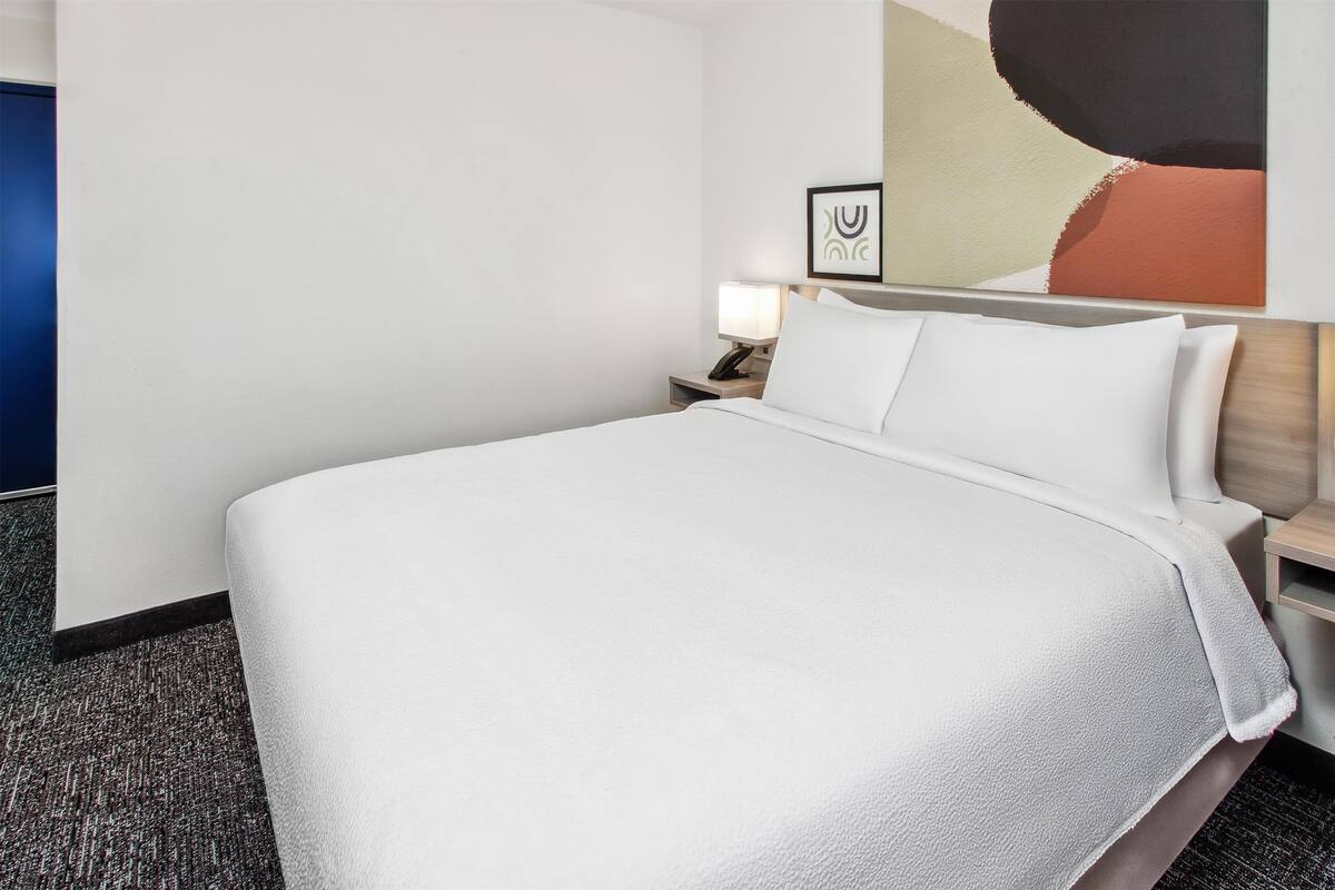 A hotel room with a large bed, a nightstand with a lamp, and a wall with abstract art.