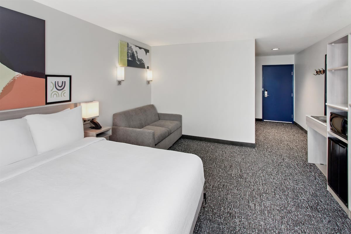 A hotel room with a large bed, a grey sofa, a desk with a lamp, and a blue door.
