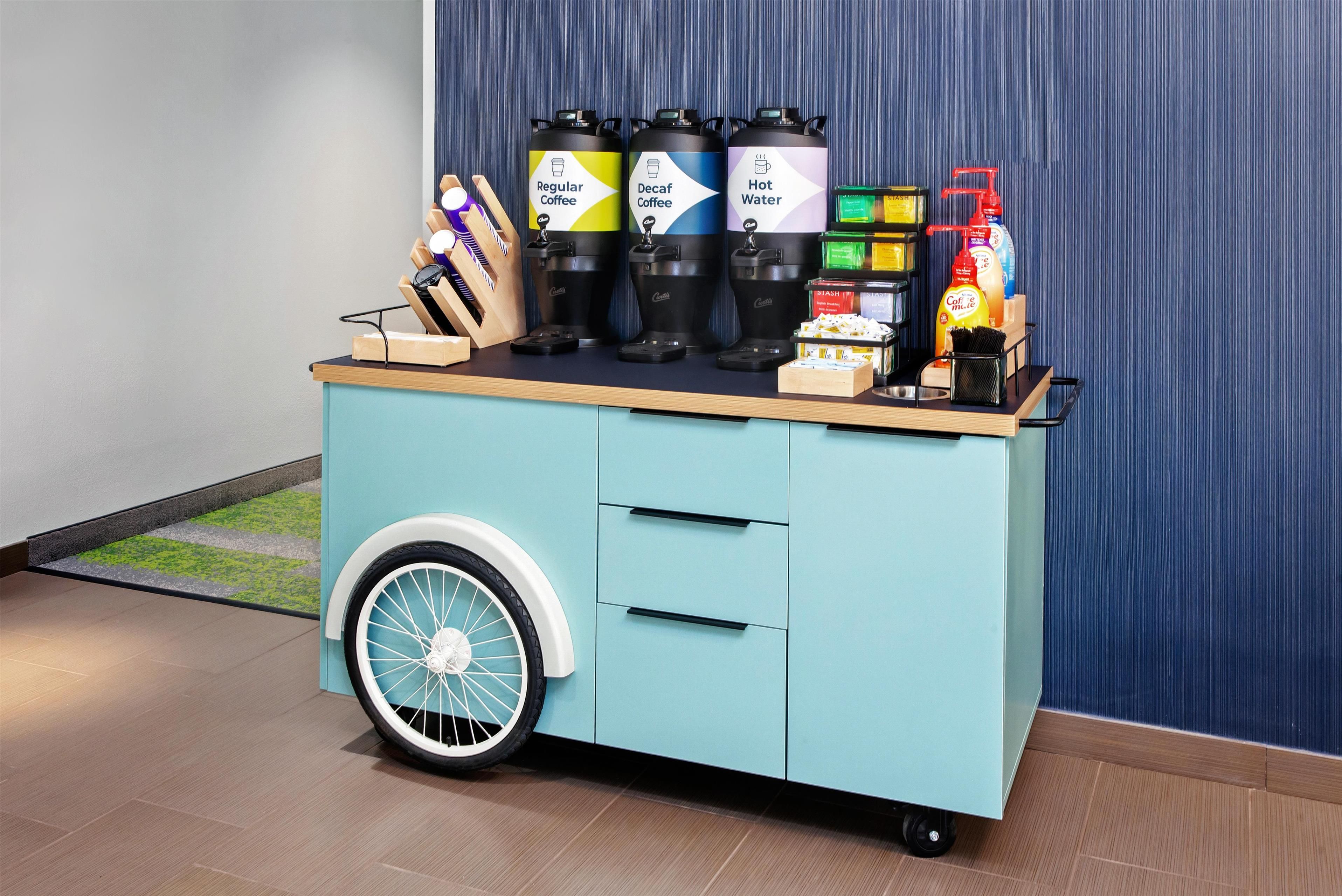A teal-colored cart with coffee dispensers, a hot water dispenser, and cleaning supplies.