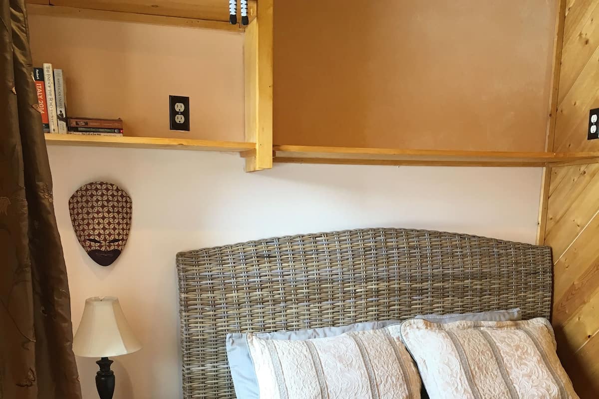 Ein Schlafzimmer mit einem gewobenen Kopfteil, einem Nachttisch mit Lampe, einem gemusterten Teppich und einer Ablage mit Büchern und Dekorationsgegenständen.