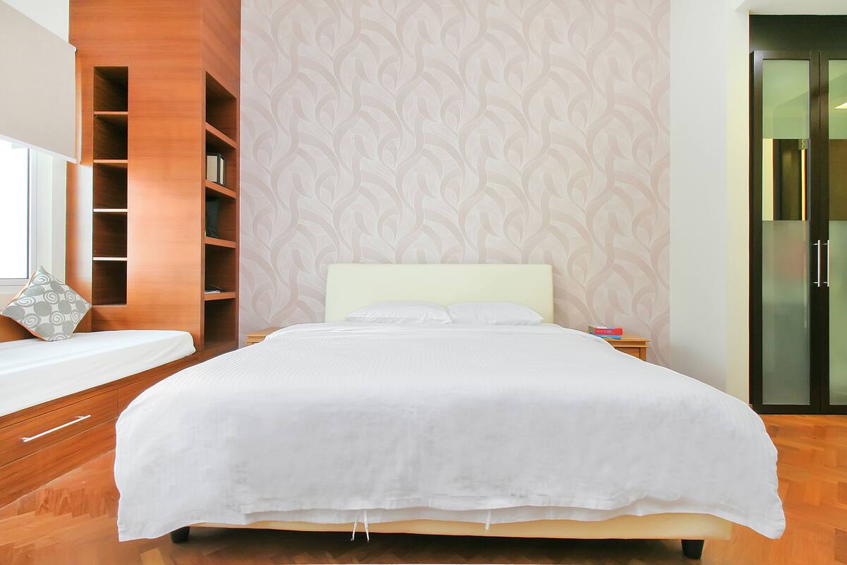 A neatly made bed with a white bedspread in a modern room with wooden flooring, a patterned wallpaper, and a built-in bookshelf.