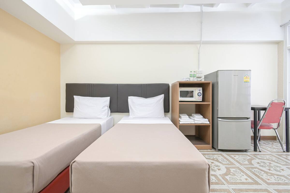 A hotel room with two single beds, a microwave, a refrigerator, a small table, and a chair.