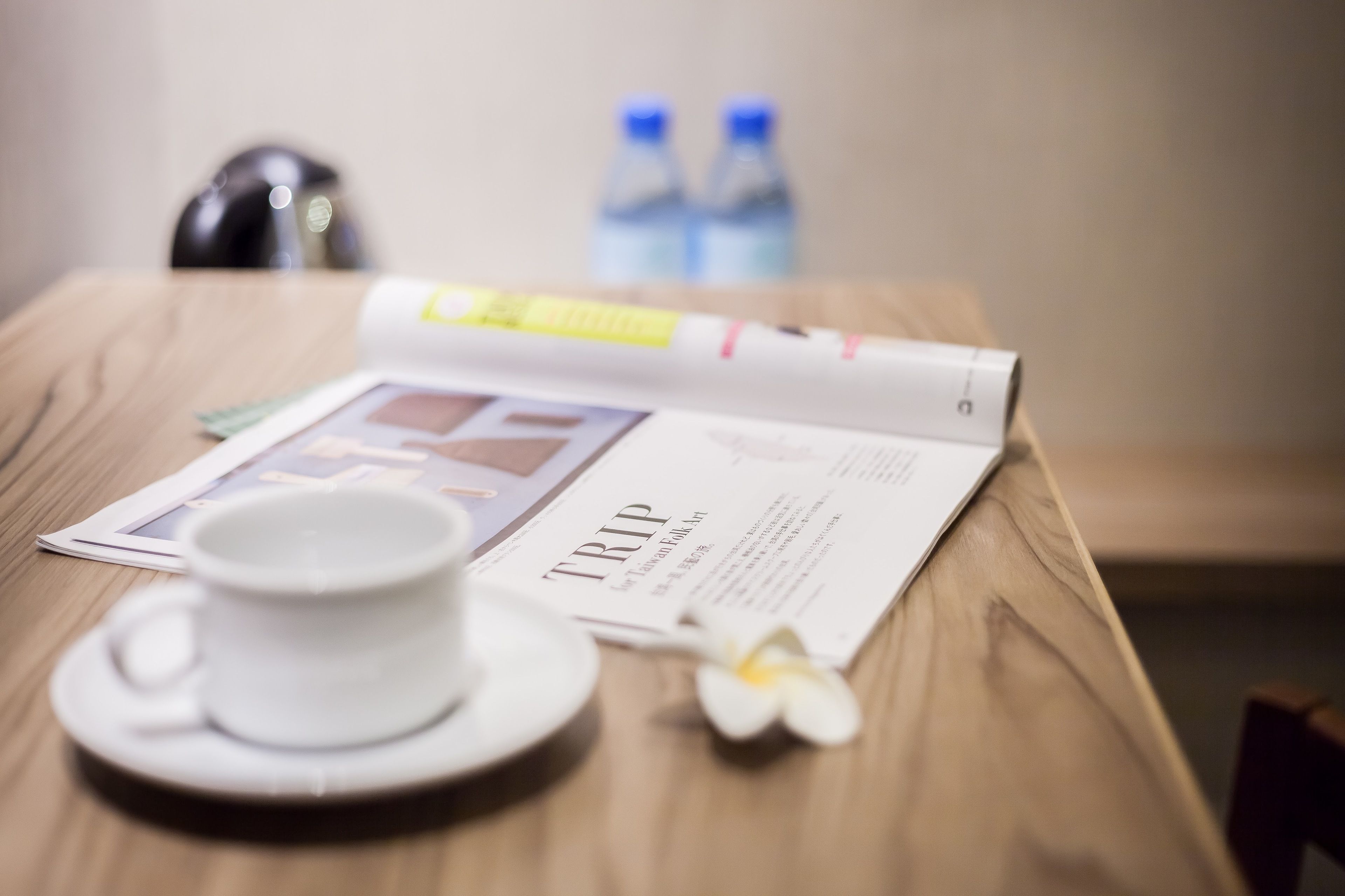 A table with a cup, saucer, a magazine, and a flower.