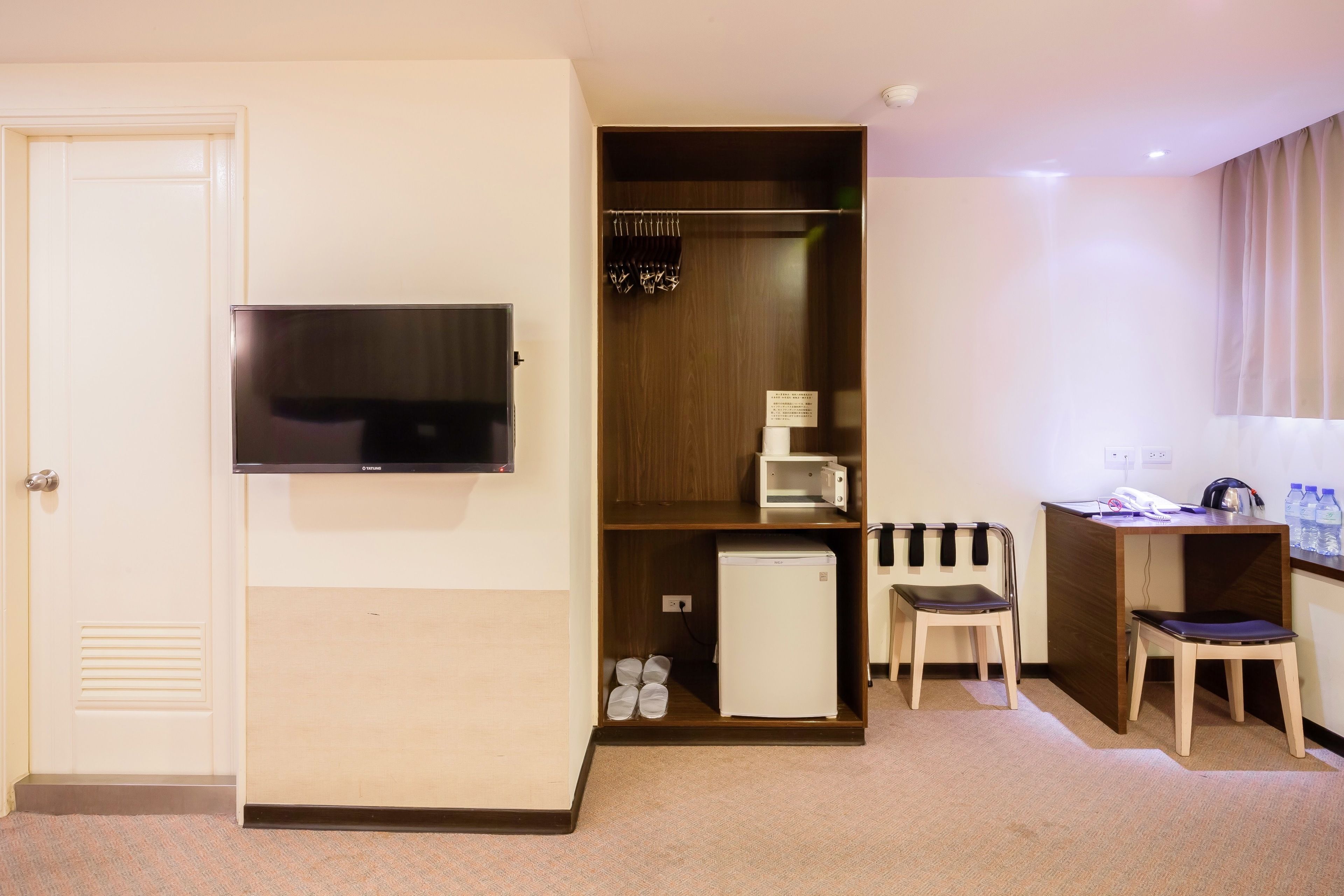 A hotel room with a television mounted on the wall, a wooden wardrobe with a mirror, a small refrigerator, a desk with a chair, and a water dispenser.
