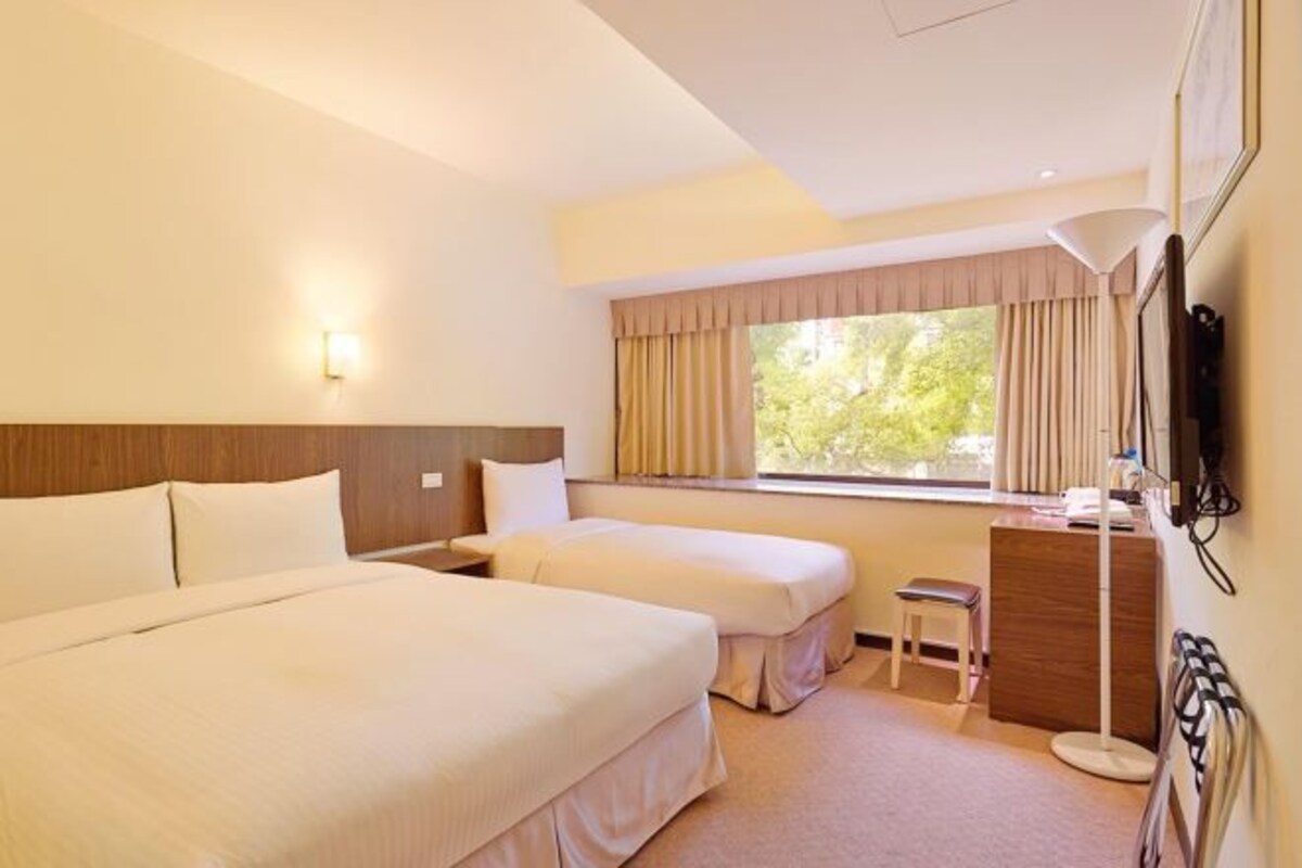 A hotel room with two beds, a desk, a chair, a standing lamp, and a window with curtains.